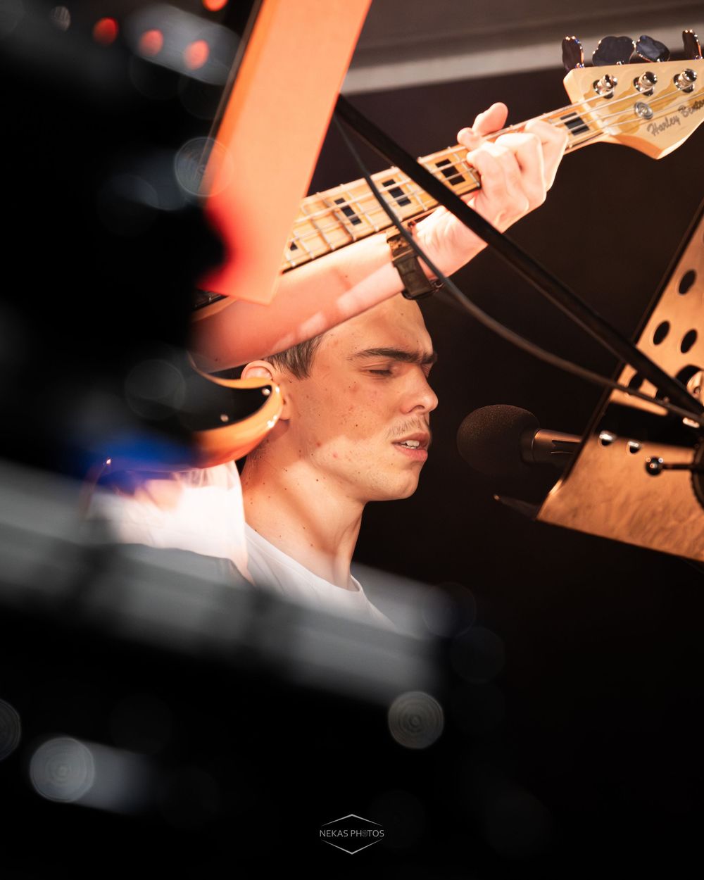 Young musician playing bass guitar on stage, captured through equipment with a microphone and warm stage lighting.