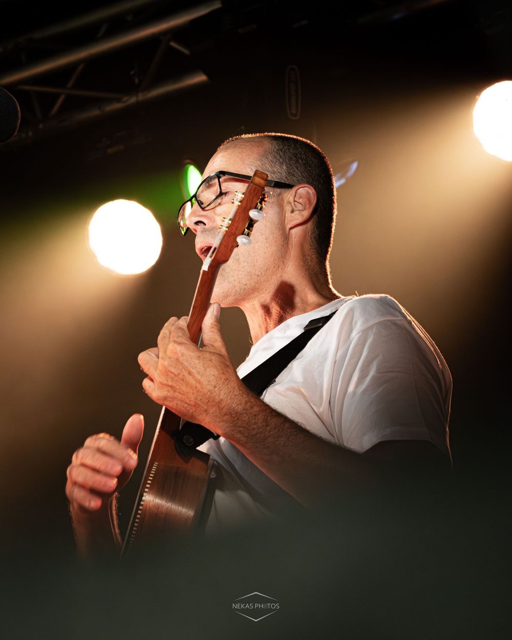 Musician playing acoustic guitar on stage under warm spotlights, wearing glasses and a white t-shirt.