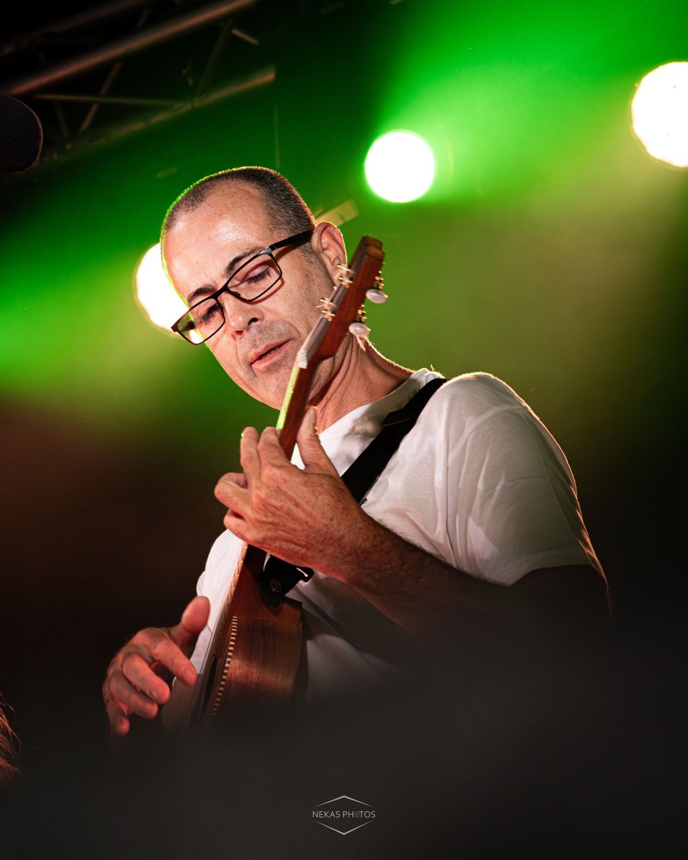 A bald guitarist with glasses performs on stage under green and white lights, playing acoustic guitar at a live concert.