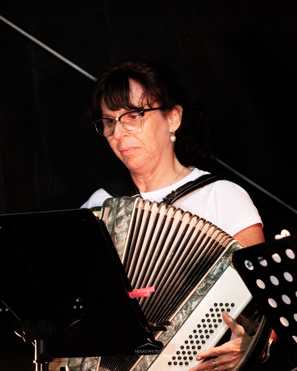 Woman with glasses playing a button accordion, reading sheet music during a live performance.