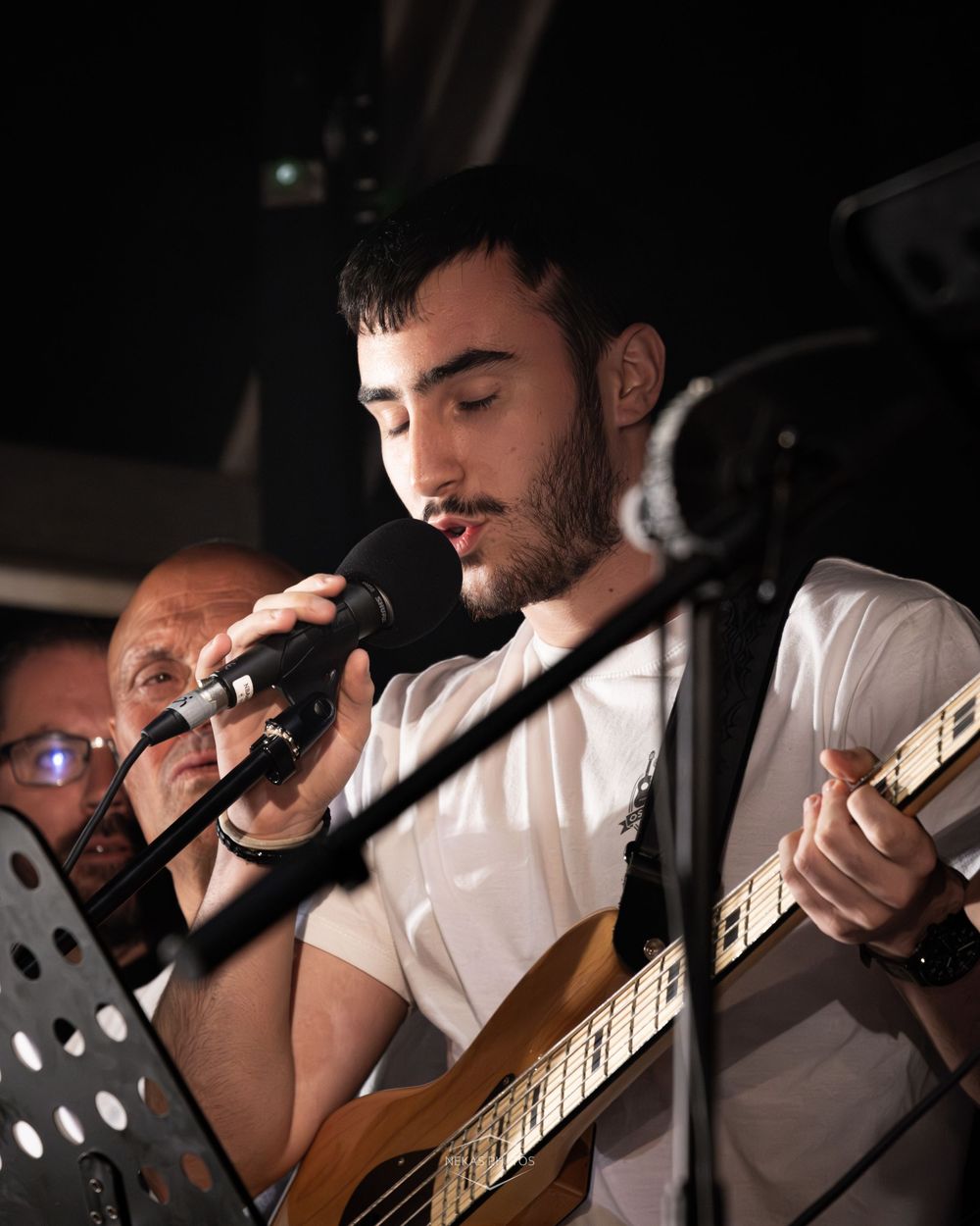A young man with stubble plays acoustic guitar and sings into a microphone on a dimly lit stage.