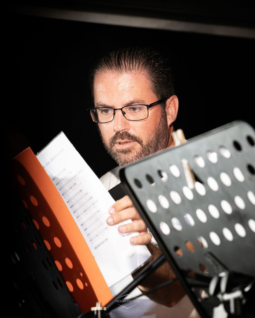 Bearded man with glasses reading sheet music at music stand during performance.