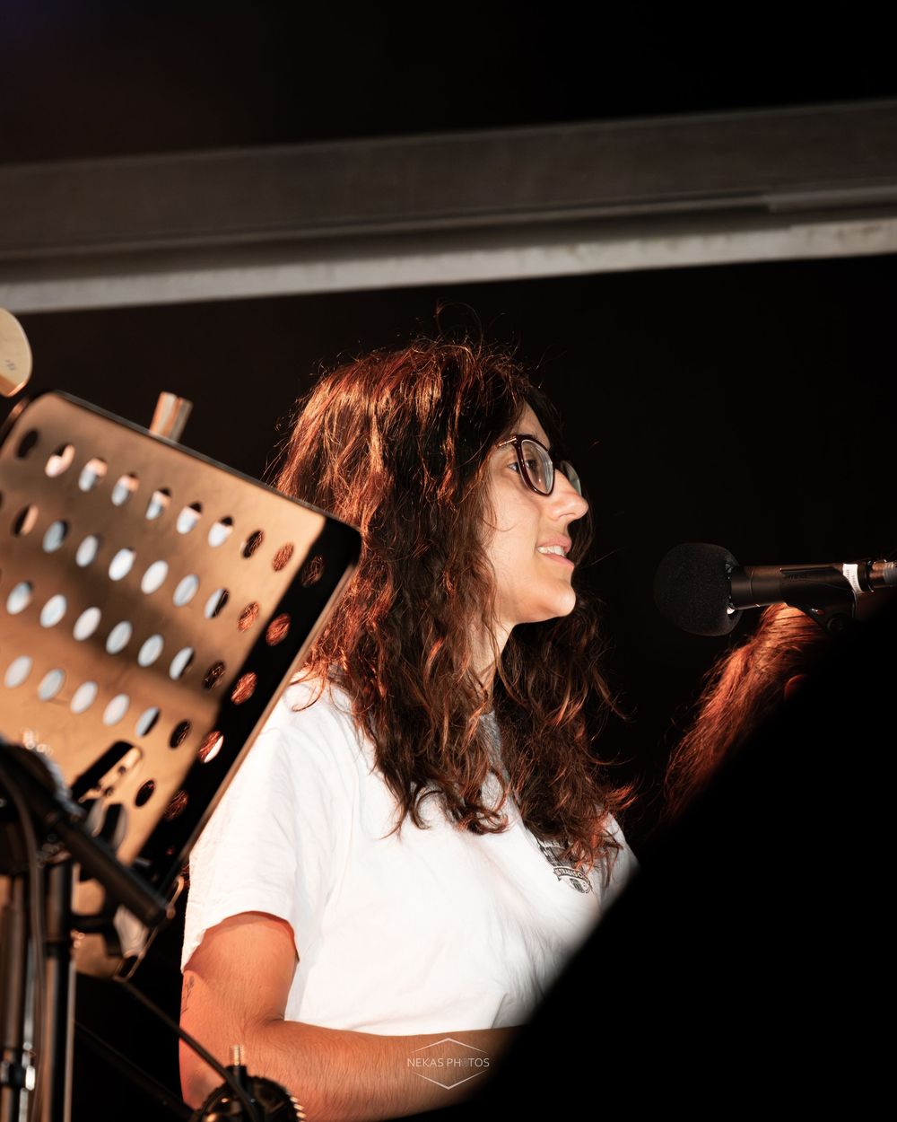 Woman with curly hair and glasses smiling while performing at microphone on stage.