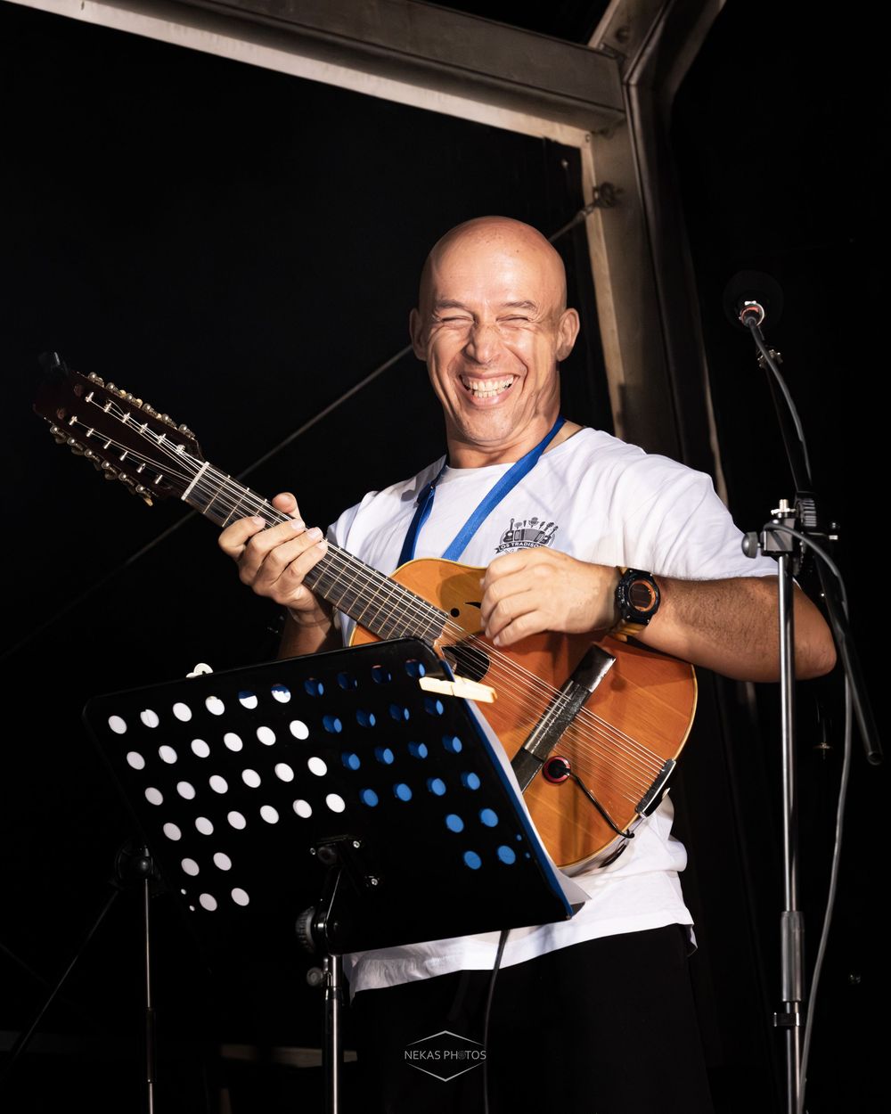 Smiling bald musician playing 12-string guitar on stage at outdoor performance event.