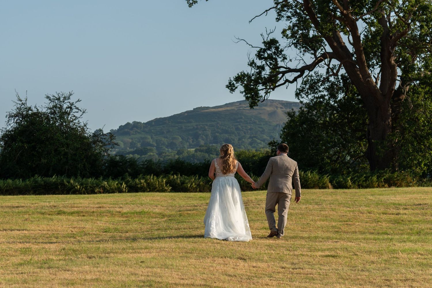 Bride and groom holding hands walking across a green field with rolling hills and trees in the background.