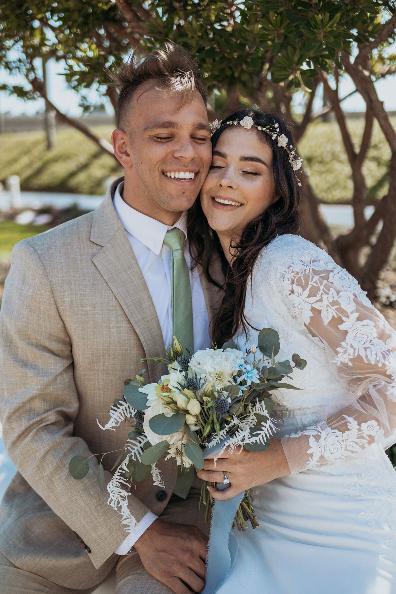 Smiling bride and groom embrace outdoors, bride holding blue and white bouquet, wearing lace dress and floral crown.