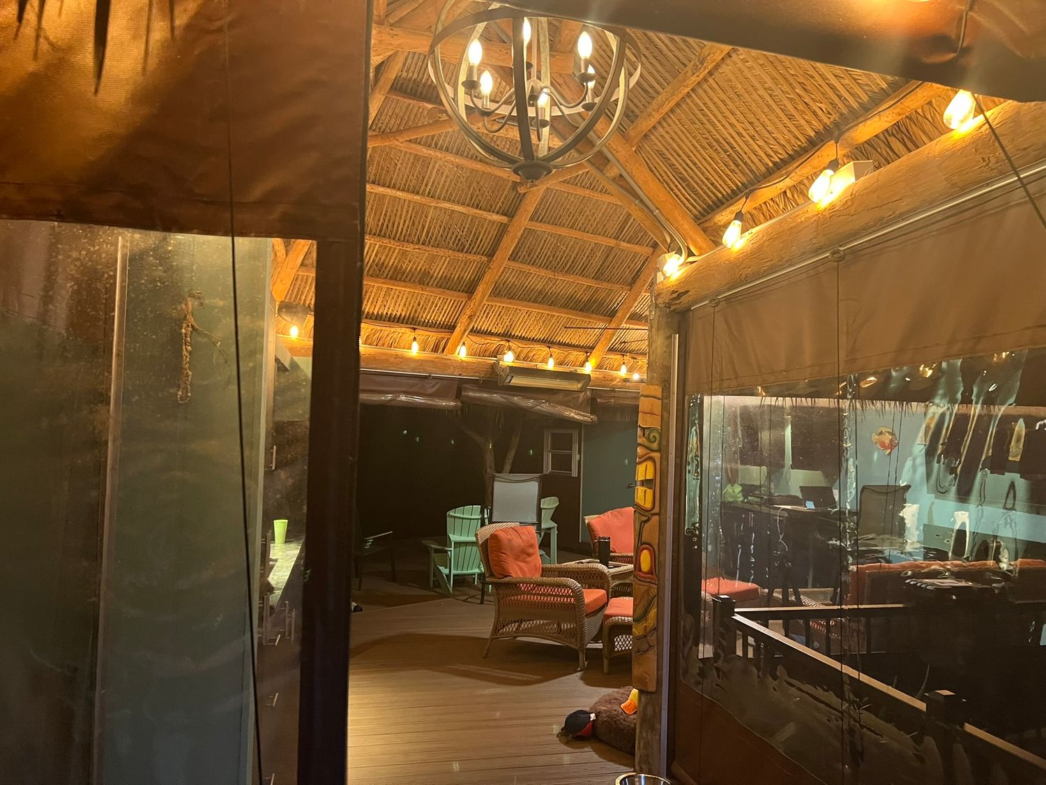 A warmly lit tiki-style covered patio with bamboo ceiling, chandelier, string lights, and comfortable seating at night.