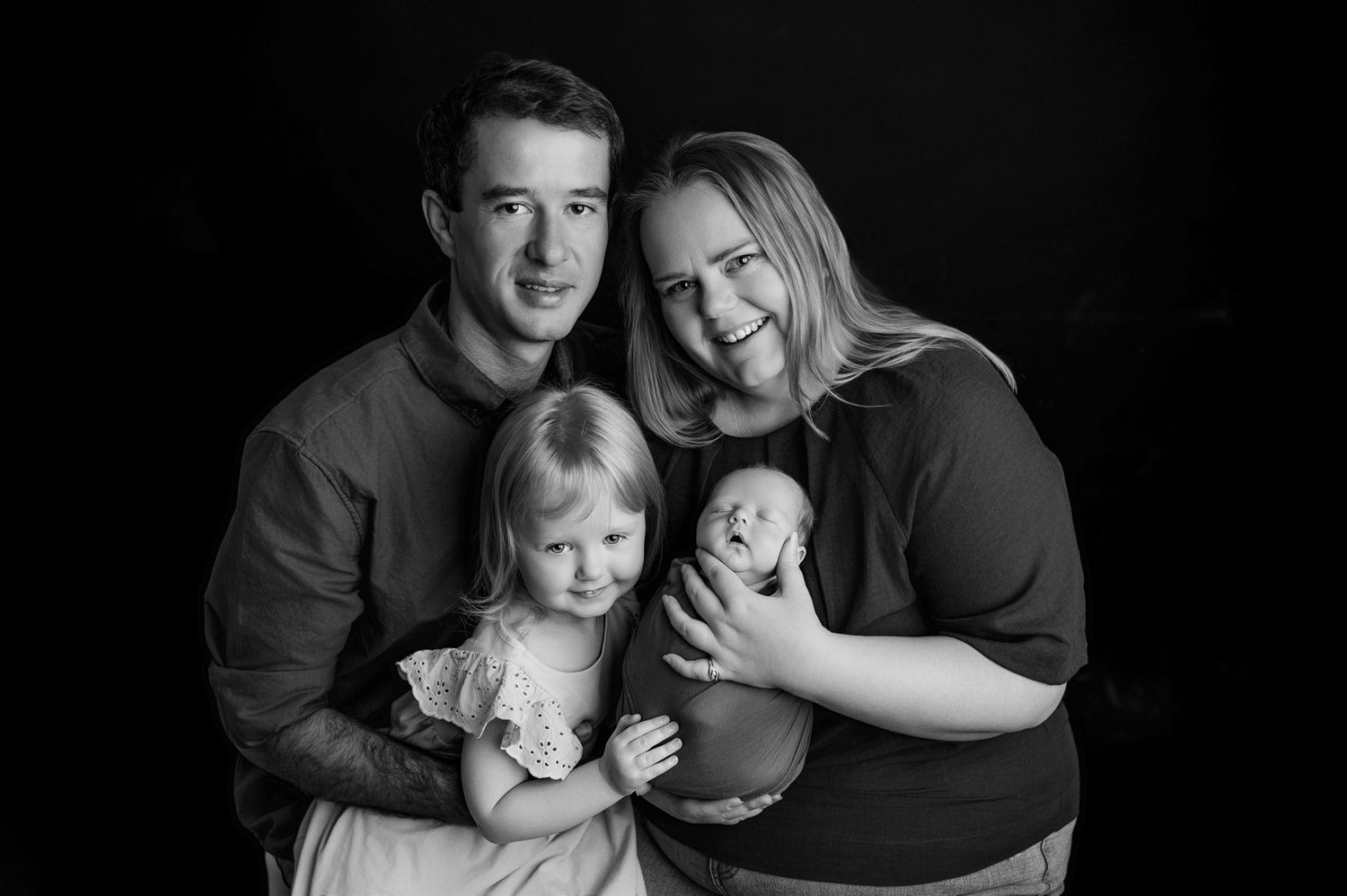 Black and white family portrait with parents and two young children, including a newborn baby, against a dark background.