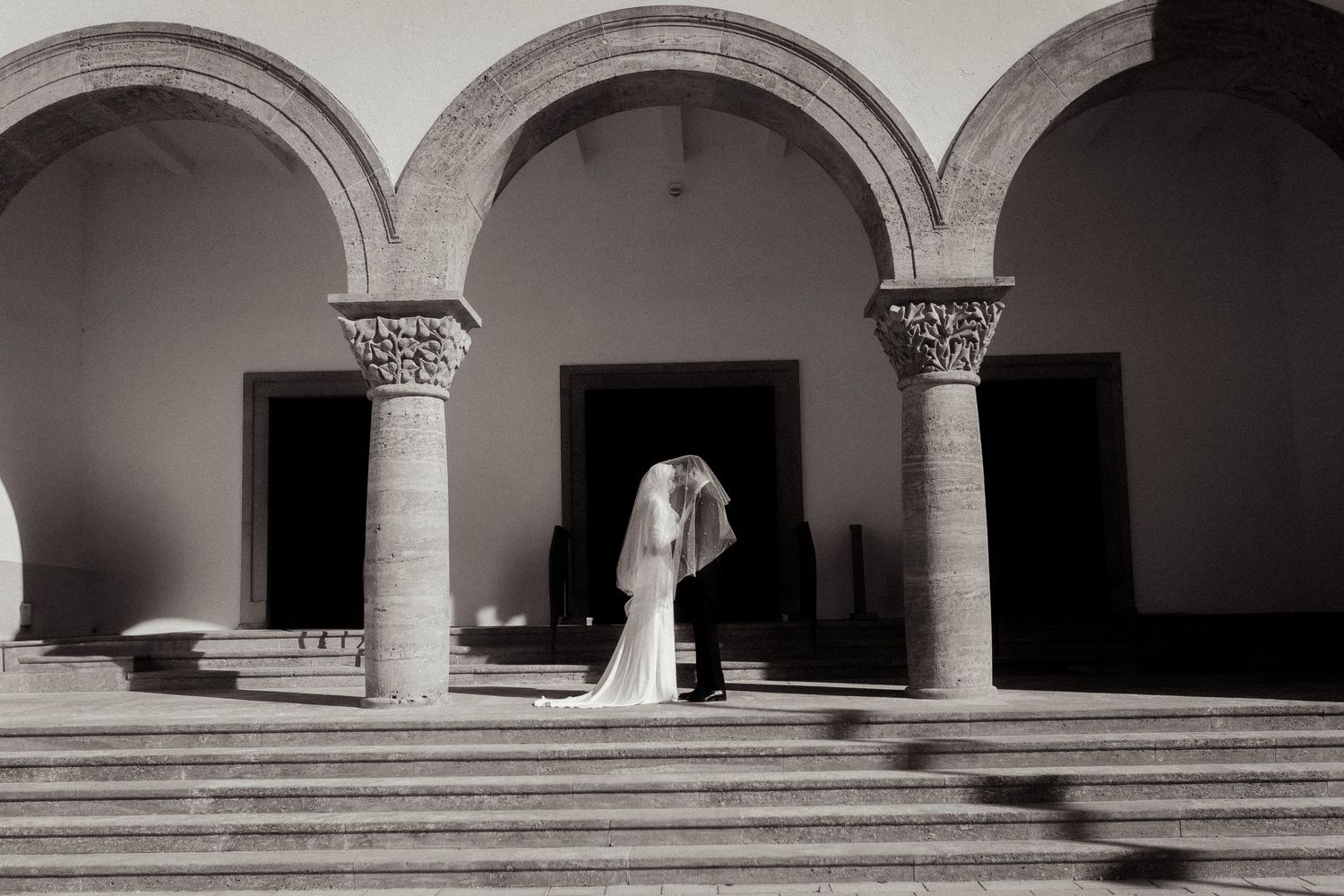Black and white photo of bride and groom kissing under elegant stone arches on steps of historic venue.