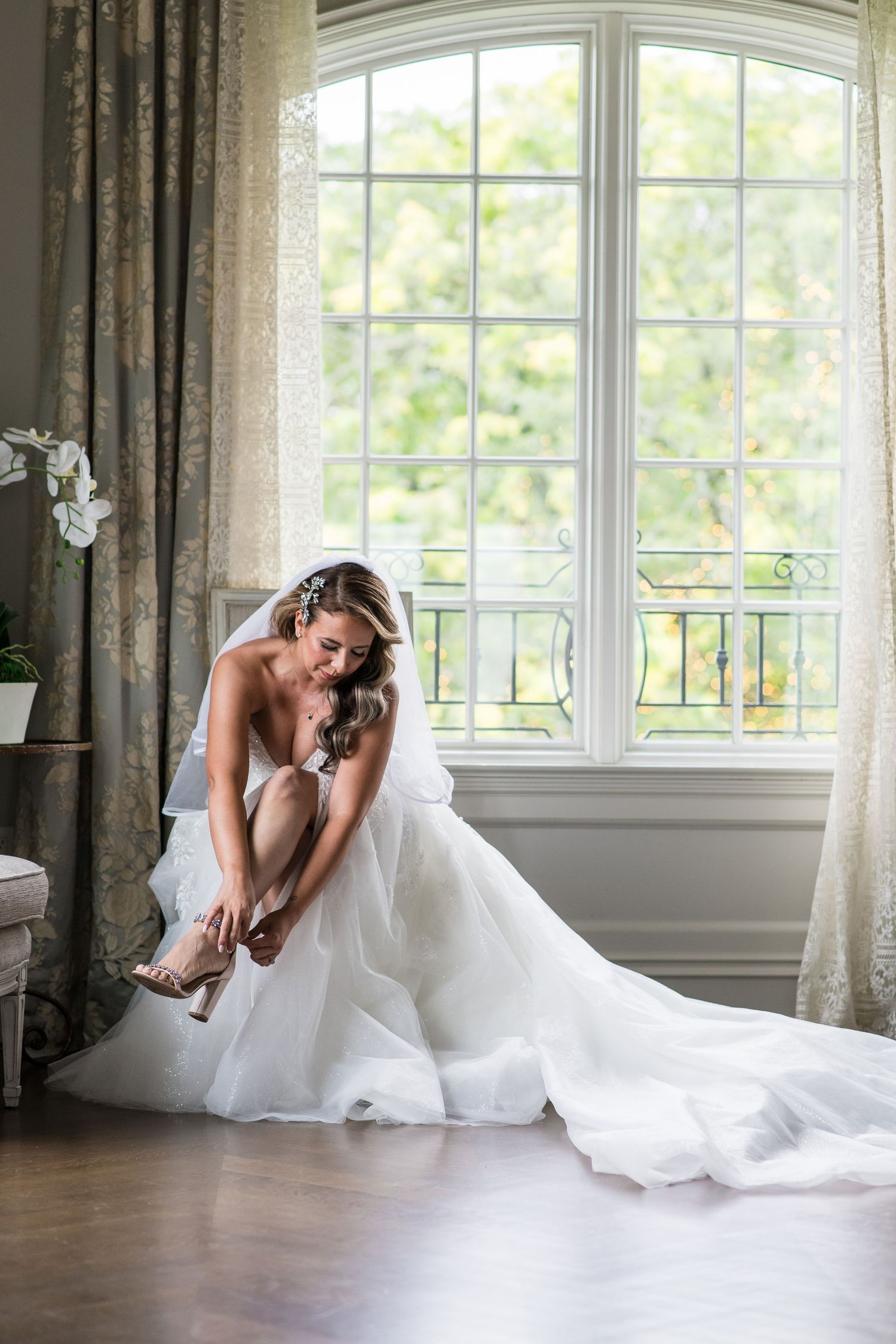 Bride in full white ballgown sitting by large window adjusting her heels in a beautifully lit bridal suite.