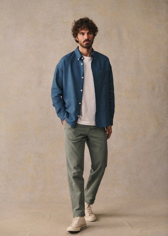 Man wearing a blue linen shirt over a white tee with sage green chinos and white sneakers.