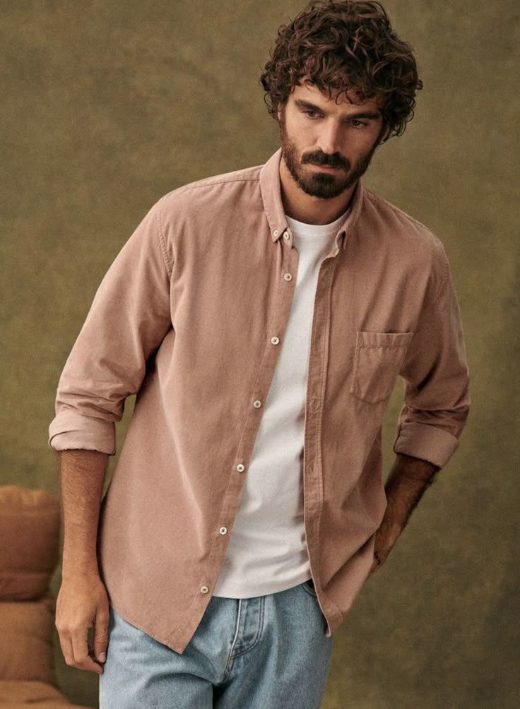 Man wearing a pink button-down shirt over a white tee with light blue jeans, styled in a casual layered look.