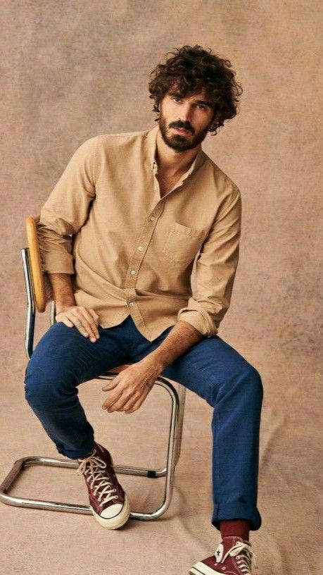 Man with curly hair and beard sits on chrome chair wearing beige shirt, blue jeans, and burgundy Converse sneakers.
