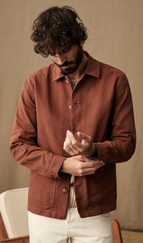 Man in rust brown chore jacket adjusting cuffs, paired with white trousers, casual layered style.