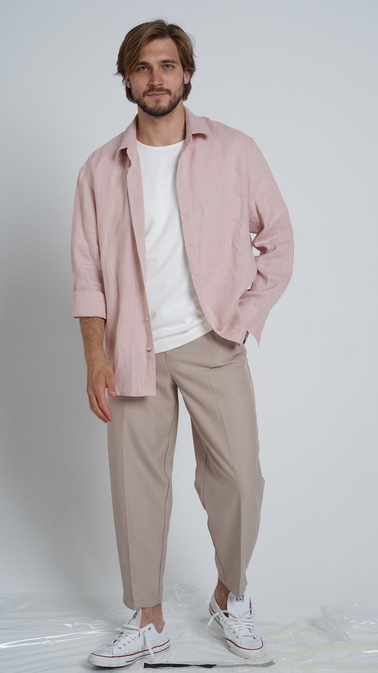 Man wearing pink linen overshirt, white tee, beige tapered trousers, and white Converse sneakers on light background.