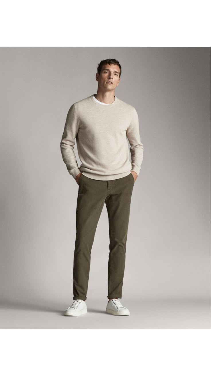 Man wearing a beige crew neck sweater, olive green chinos, and white sneakers against a grey background.