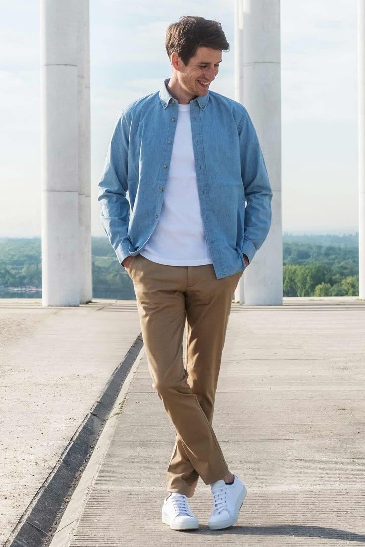 Young man in blue shirt, white tee, khaki pants, and white sneakers walking outdoors near white columns.