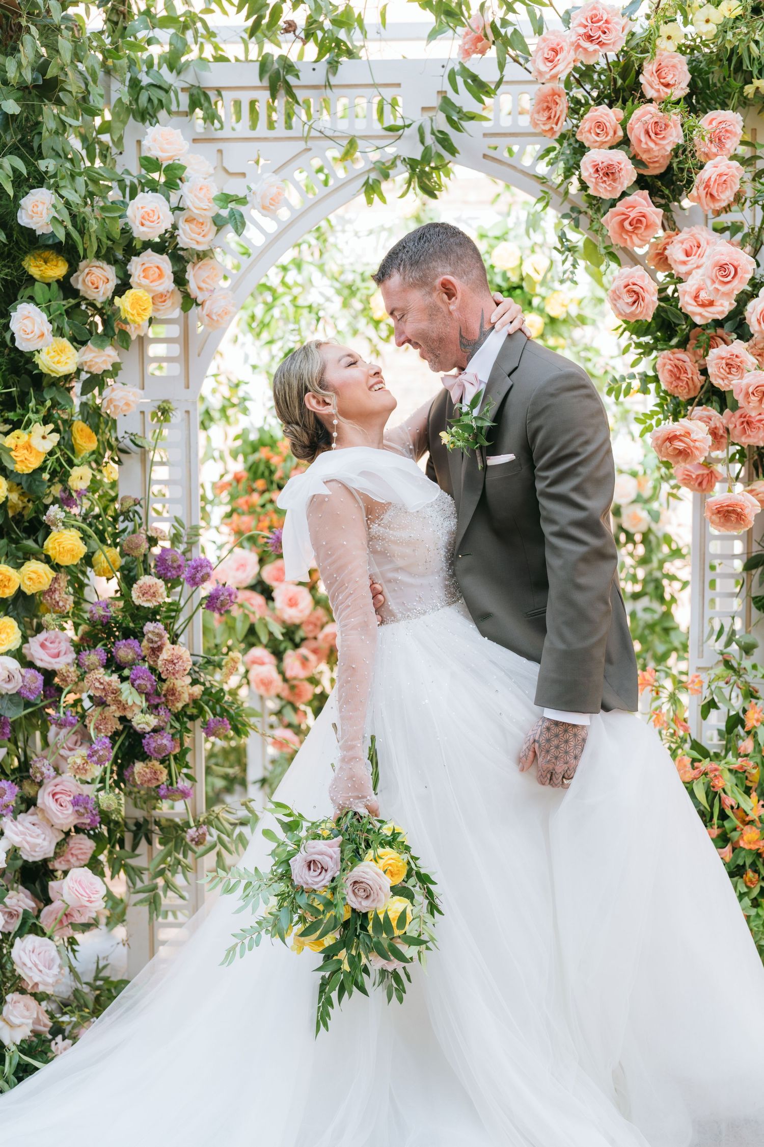 Bride in white ball gown and groom in black tuxedo posing before a vine-covered white building with stained glass windows.