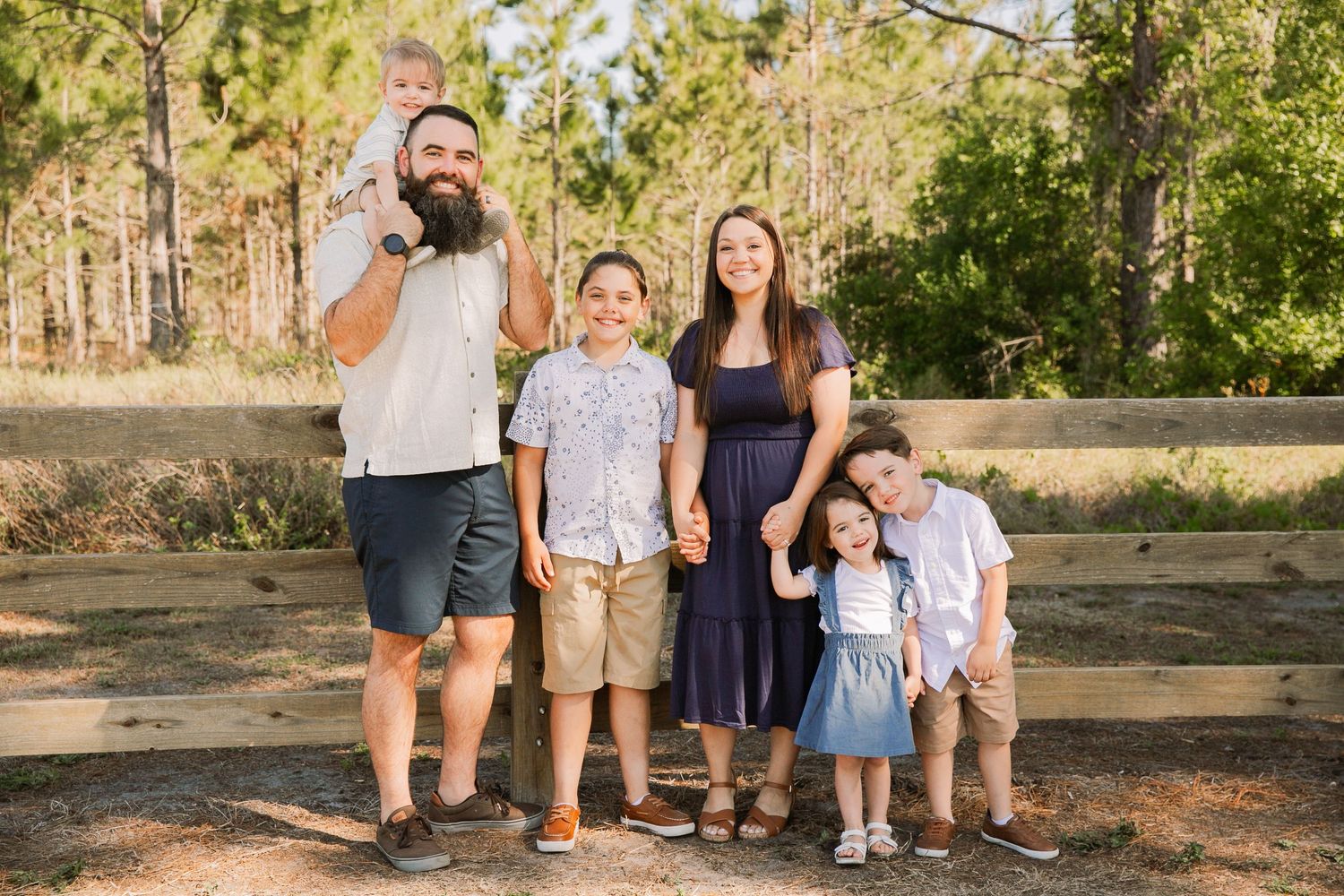 Family of six laughing and embracing outdoors in casual blue outfits during a warm sunny day photo session.