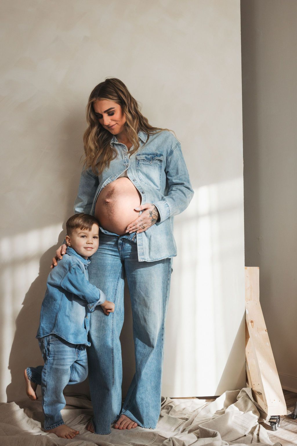 Pregnant woman in denim outfit with toddler in matching denim, posing together in a bright, minimalist room.