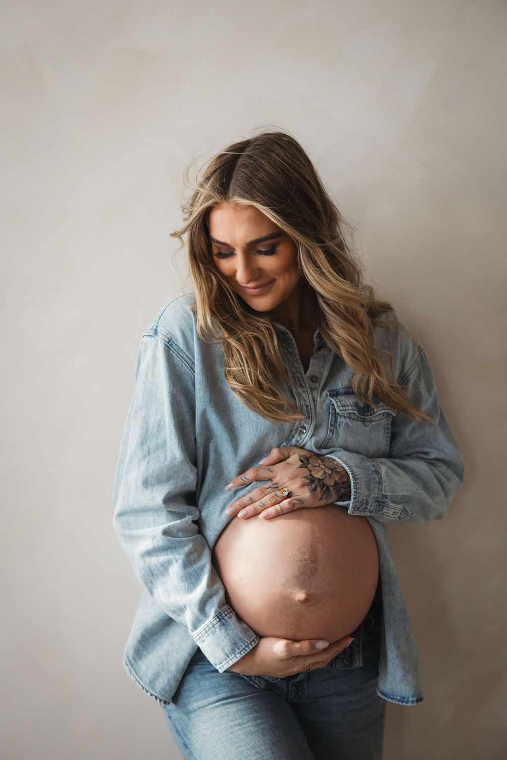 Smiling pregnant woman in denim shirt cradling her bare baby bump, with long wavy hair and a hand tattoo.