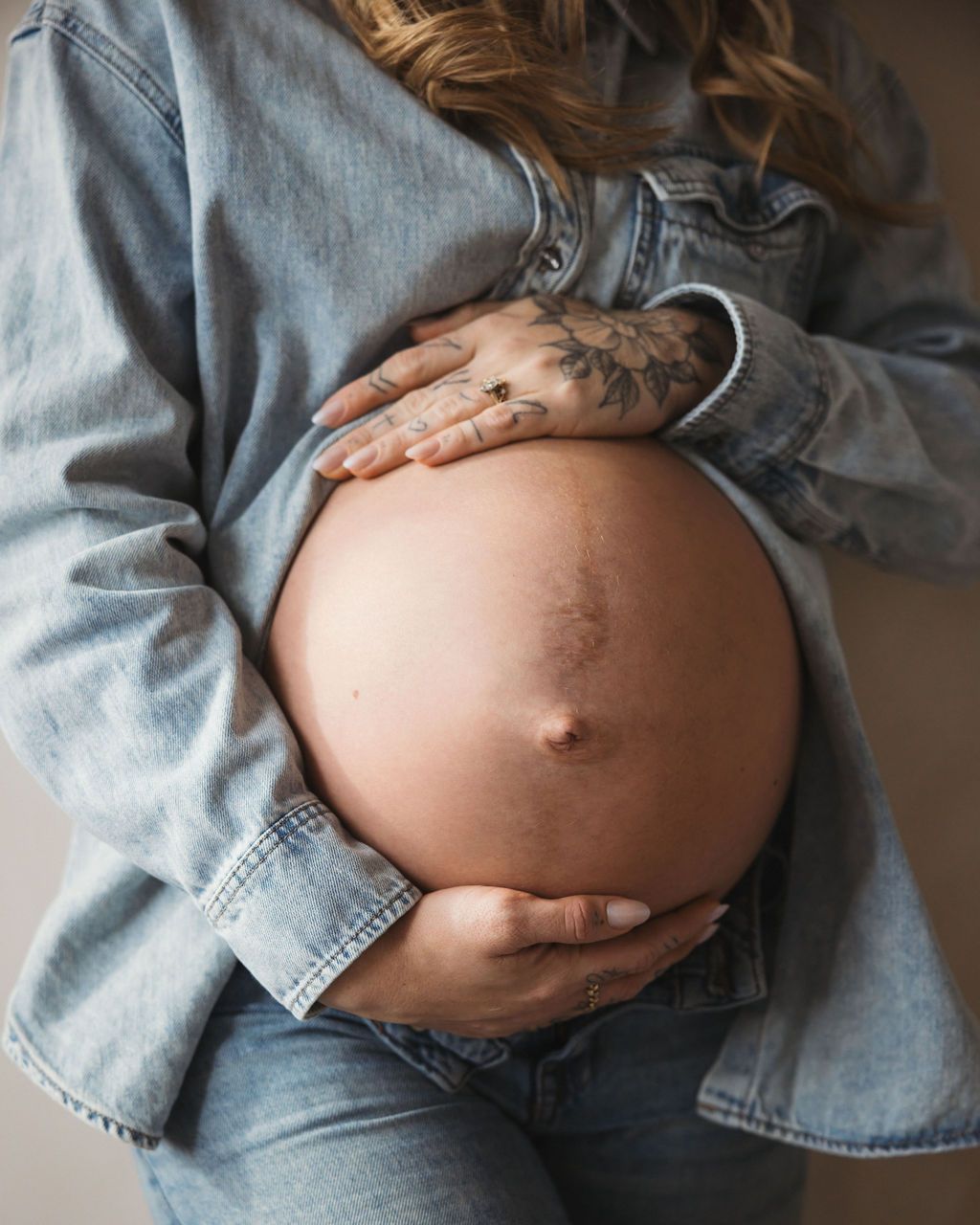 Pregnant woman in denim jacket cradling bare baby bump with tattooed hands, maternity photography.