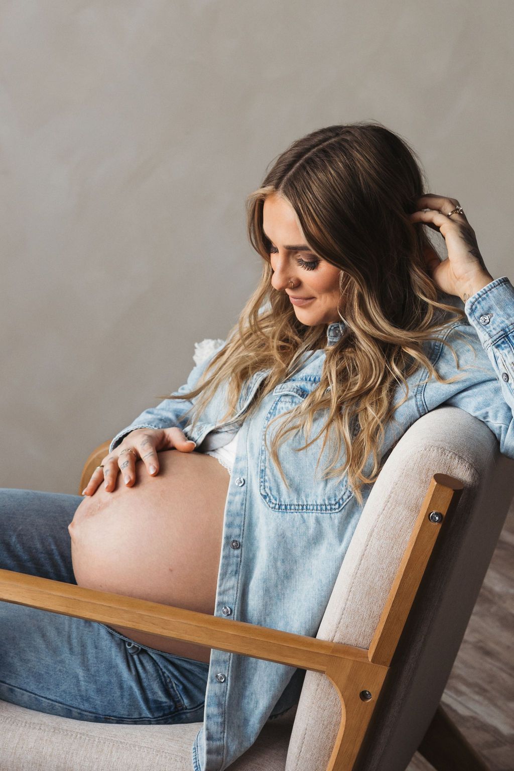 Pregnant woman in denim shirt sitting in chair, gently touching her baby bump and smiling downward.