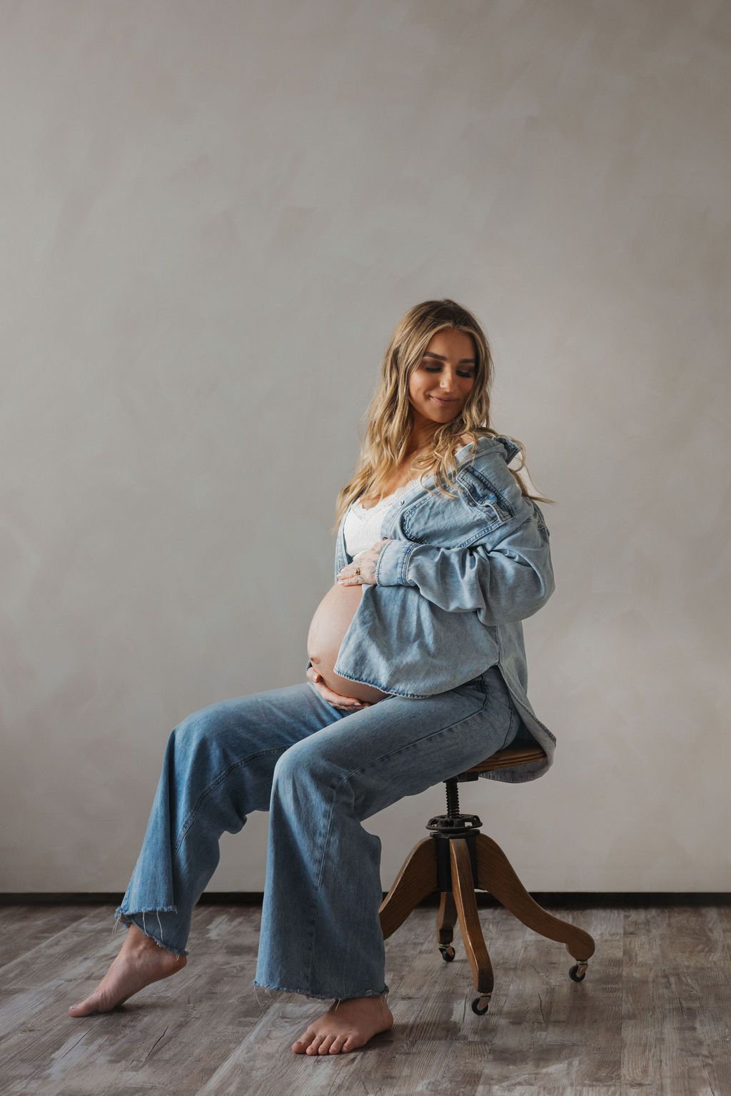 Pregnant woman in denim jacket and jeans sits on chair cradling baby bump in a studio setting.