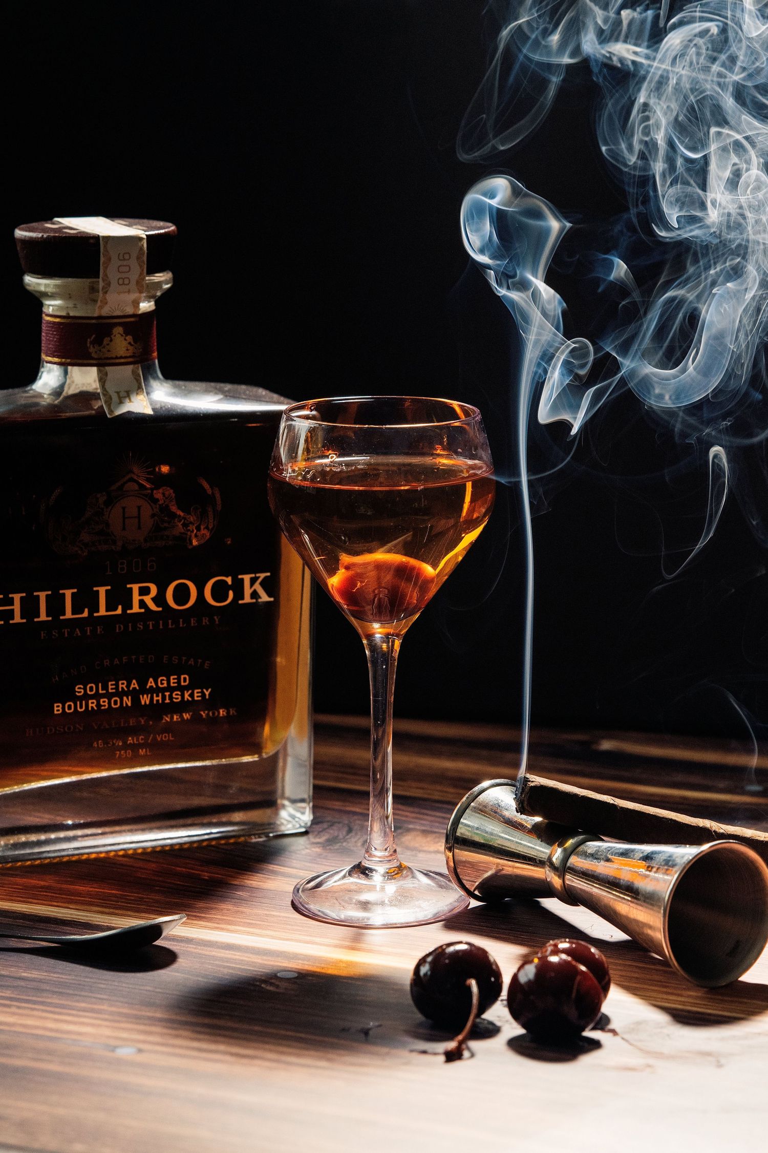 Hillrock Solera Aged Bourbon Whiskey bottle beside a cocktail glass with smoke rising, cherries, and a jigger on wood.