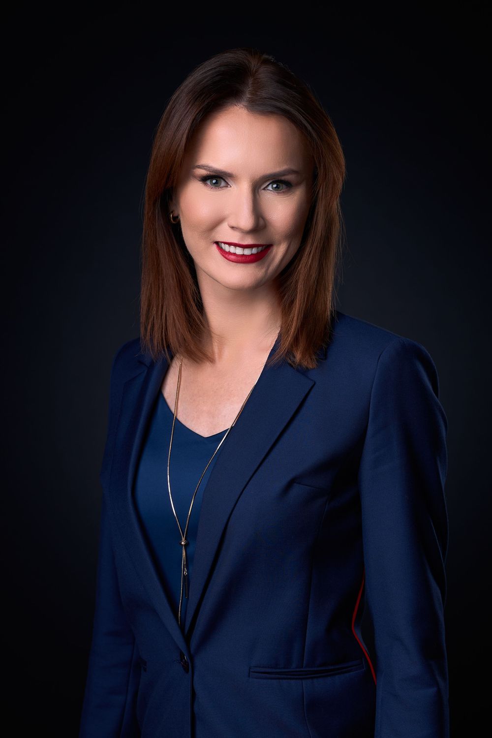 Professional woman in navy blazer with red lipstick and necklace smiling confidently against dark background.
