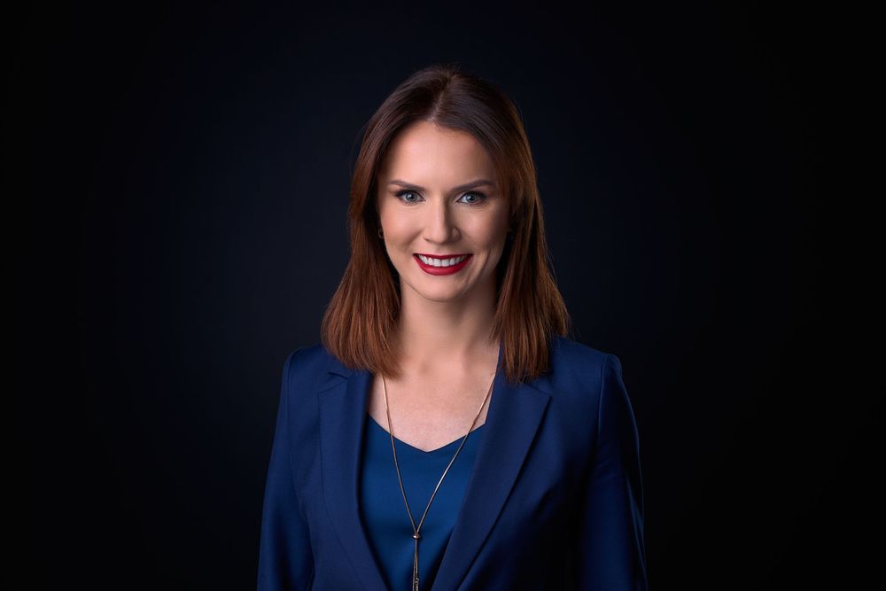 Professional woman in blue blazer smiling confidently against dark background, corporate headshot portrait.