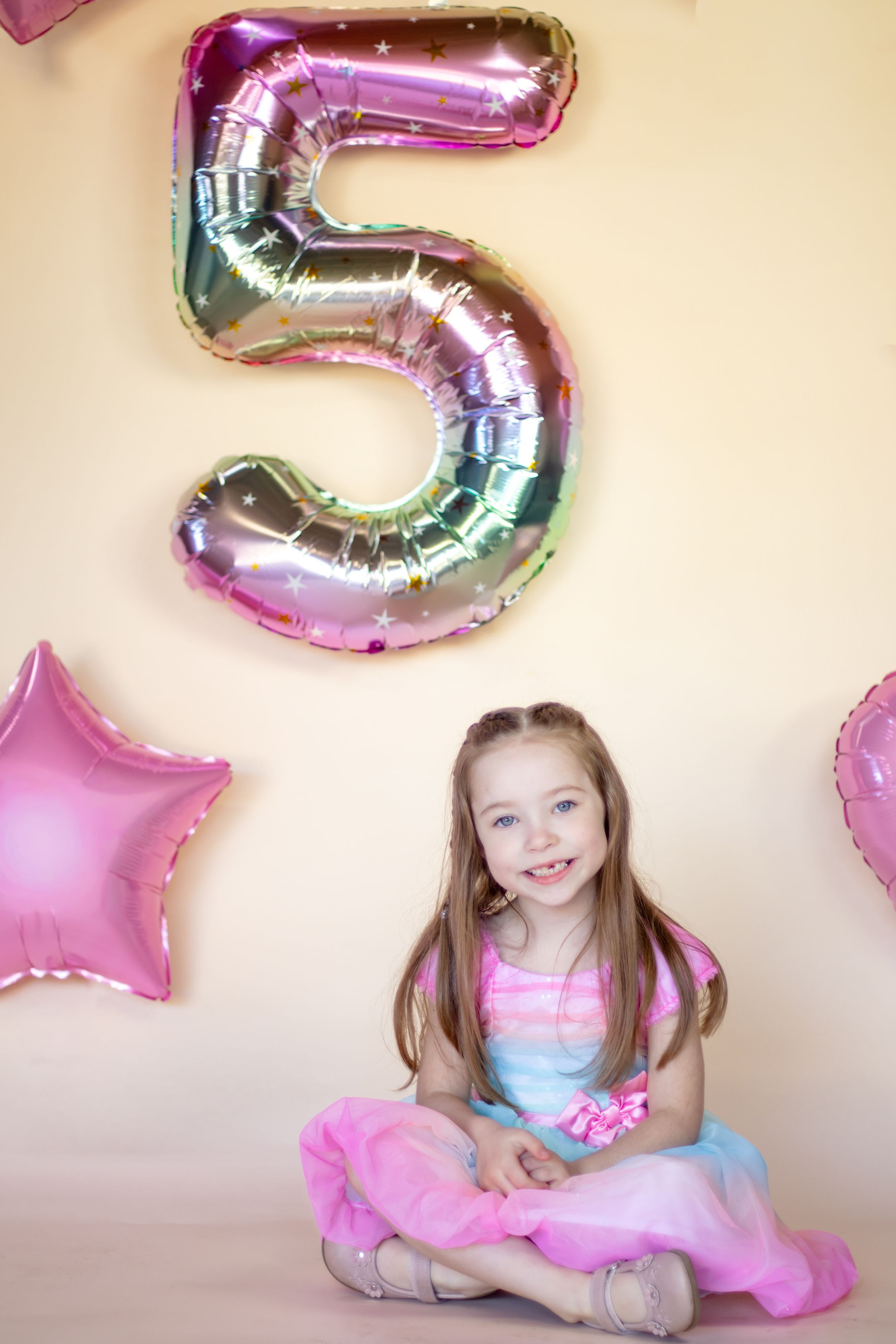A smiling girl in a pink tutu sits cross-legged in front of a large number 5 balloon for her 5th birthday photoshoot in washington studio