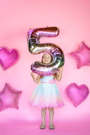 A young girl in a colorful dress holds a large number 5 foil balloon surrounded by pink heart and star balloons taken in roy wa studio