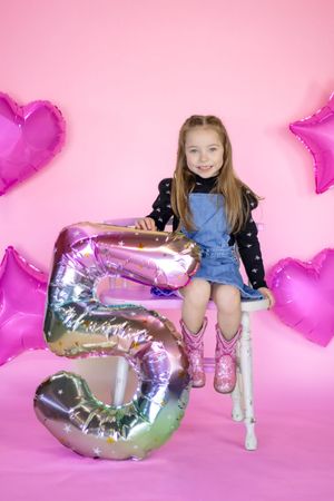 A smiling girl in a denim dress and pink cowboy boots sits beside a large number 5 balloon in pink studio setting in roy washington
