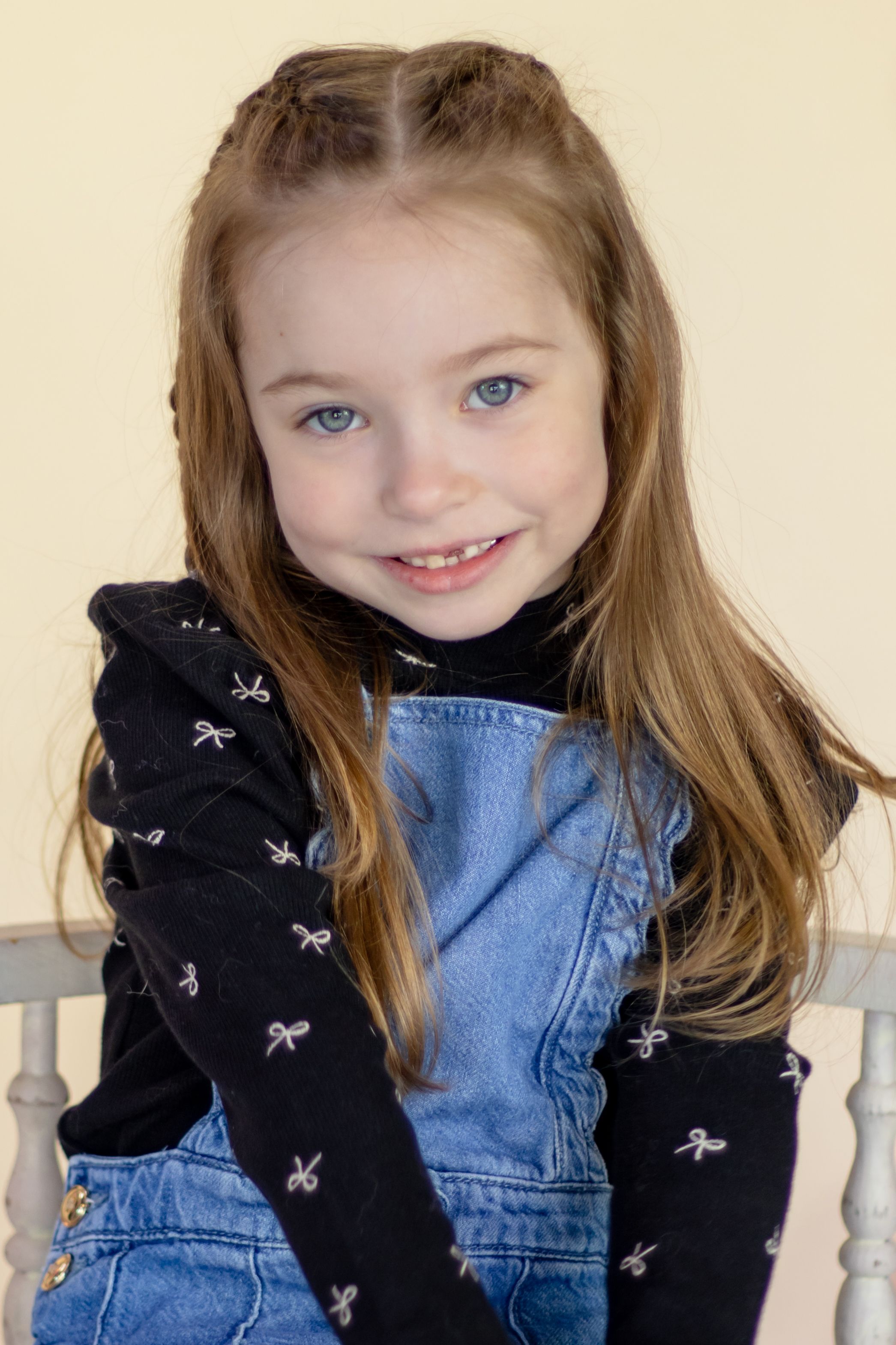 A cute young girl with blue eyes and long brown hair smiles wearing a denim jumper and black bow-print top taken by washington photographer