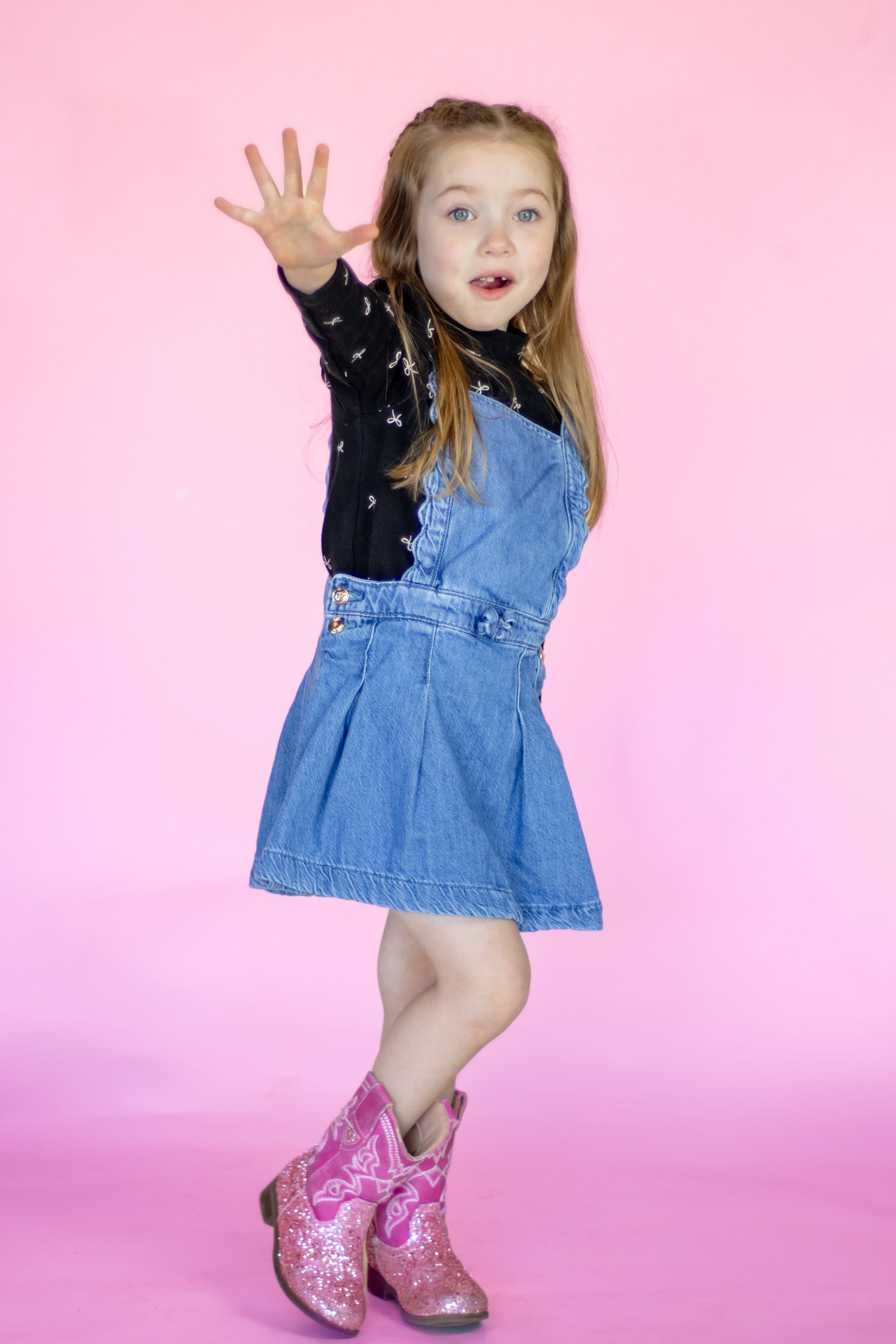 A playful young girl in a denim skirt and pink cowboy boots poses with her hand raised on a pink background in roy washington studio
