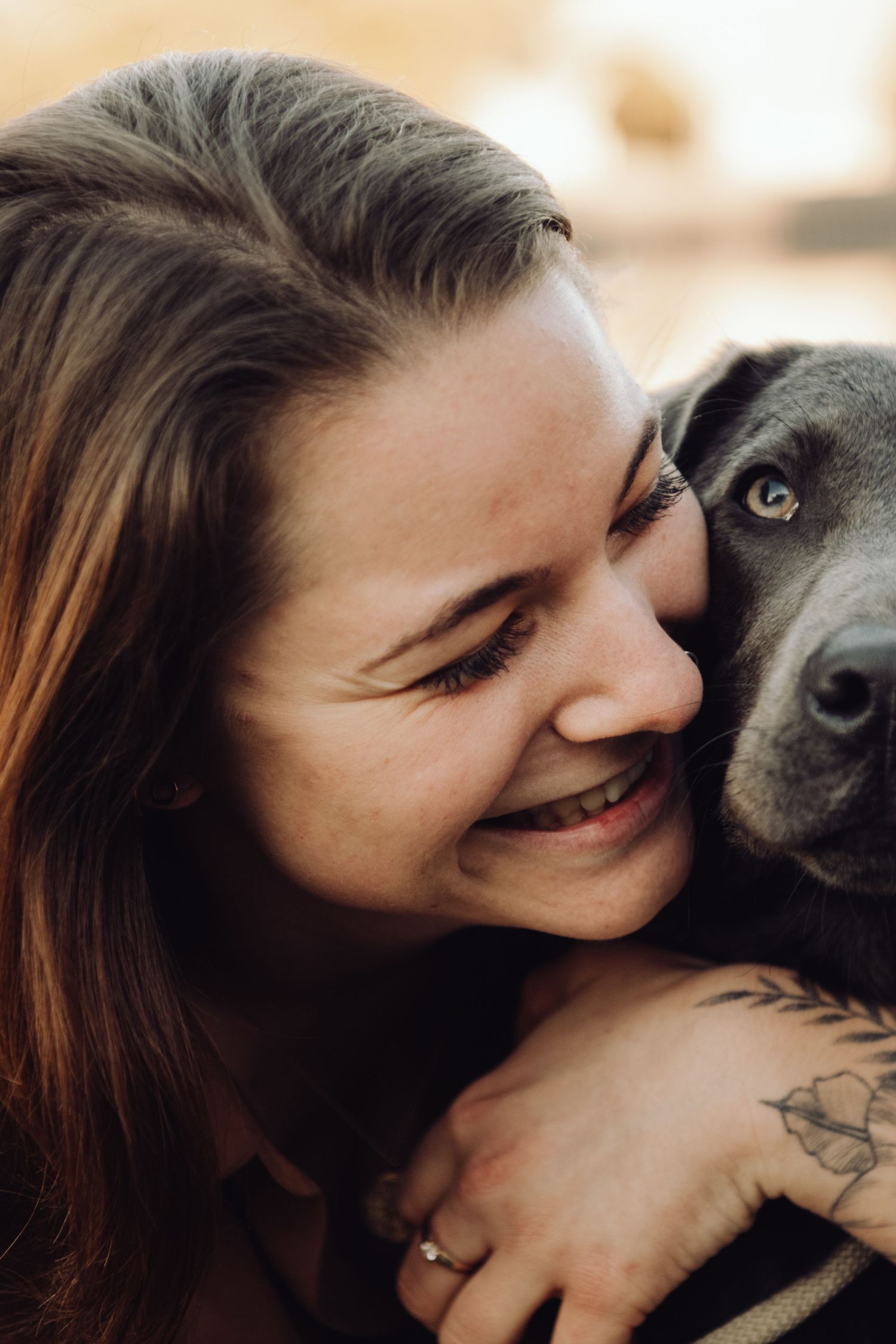 A smiling woman with a tattoo hugs a black Labrador dog closely, sharing a joyful moment together outdoors.