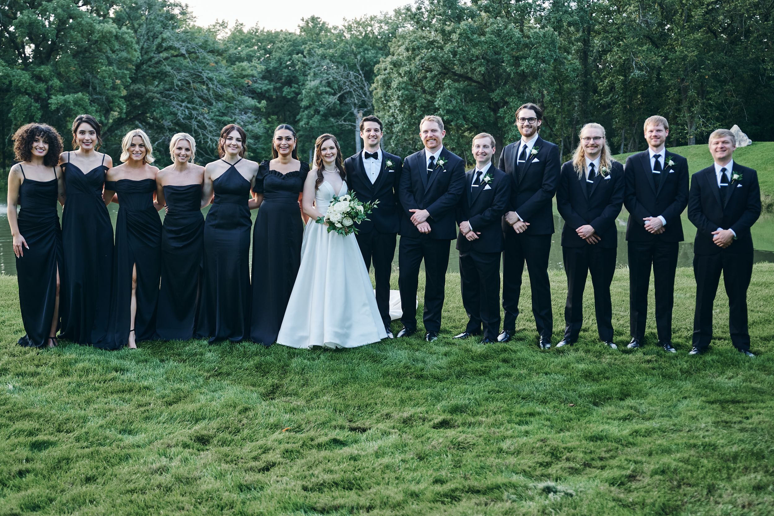 Wedding party in black attire posing outdoors on green grass, bride in white gown holding bouquet at center.