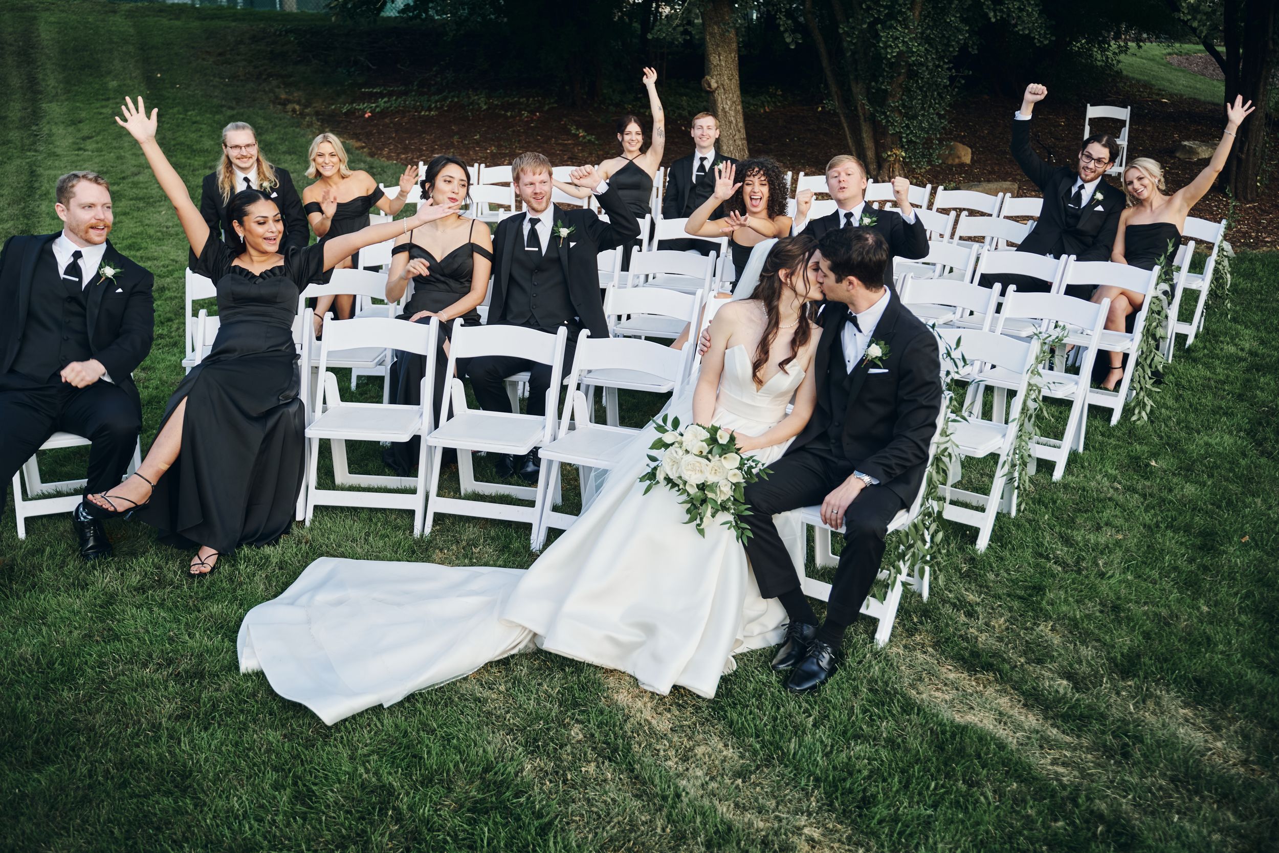 Bride and groom share a kiss at outdoor wedding ceremony while guests in black formal attire cheer and celebrate behind them.