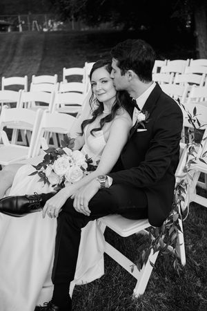 Black and white photo of a bride and groom sitting in white chairs at an outdoor wedding ceremony, sharing a tender moment.