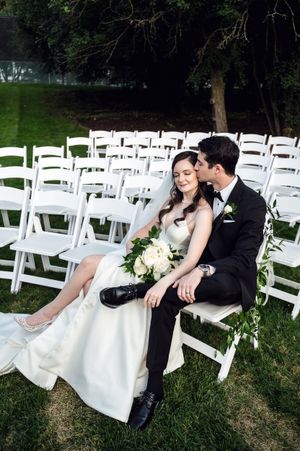 Bride and groom sit together among white chairs at outdoor wedding ceremony, sharing a tender kiss.