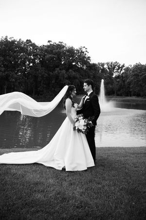 Black and white photo of bride and groom standing by a pond with a fountain, veil flowing in the wind.