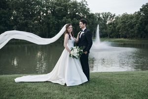 Bride and groom pose by a serene pond with a fountain, her veil flowing gracefully in the wind on their wedding day.