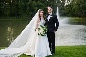 Bride and groom posing together outdoors near a pond with a fountain, bride wearing a white gown with long veil.