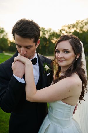 Groom in black tuxedo kissing bride's hands while she smiles softly, outdoor wedding portrait at sunset.