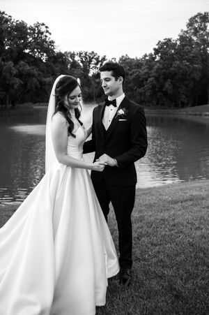 Black and white photo of a bride and groom holding hands by a serene pond, surrounded by lush trees.