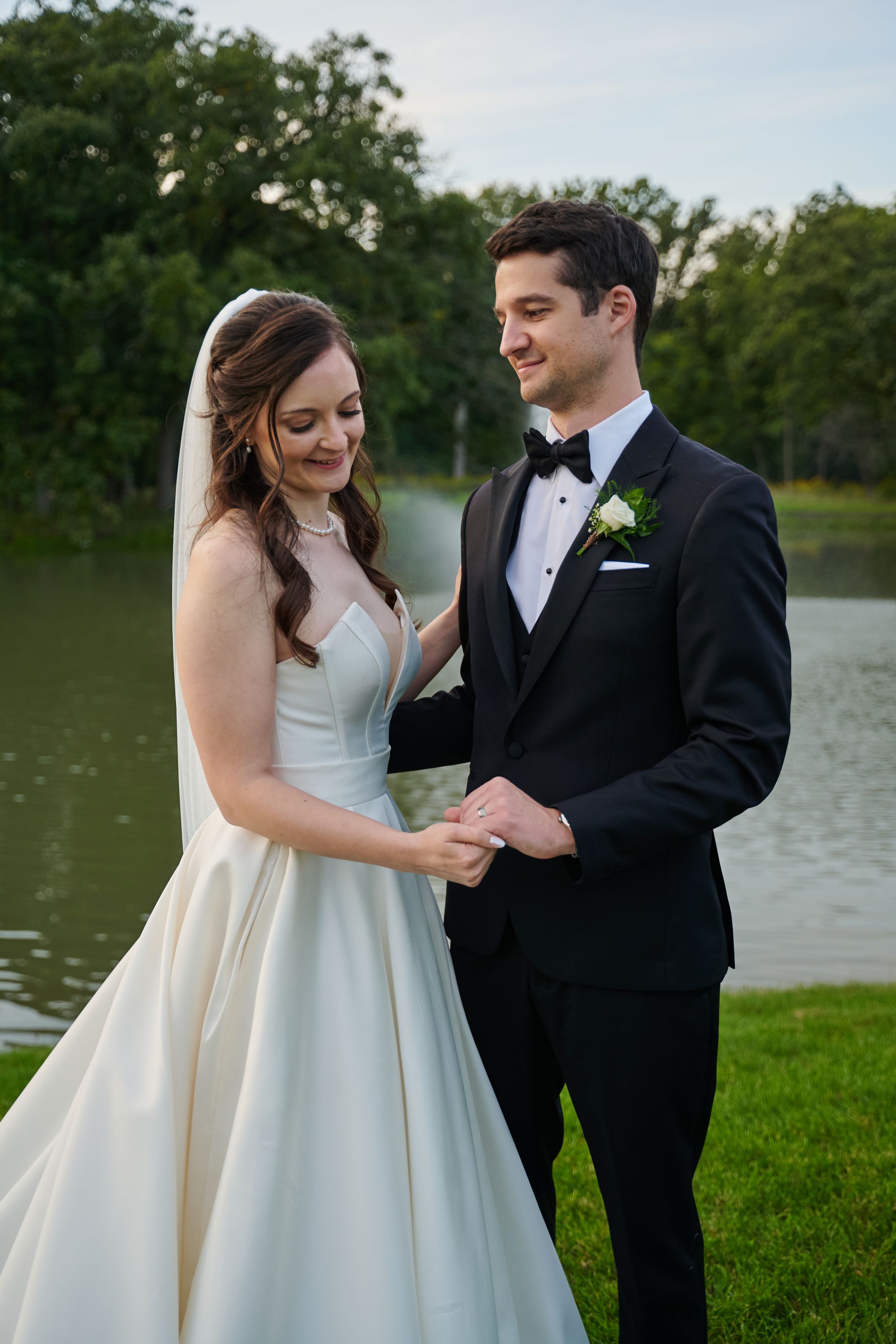 A bride and groom hold hands near a serene pond, smiling warmly on their wedding day outdoors.