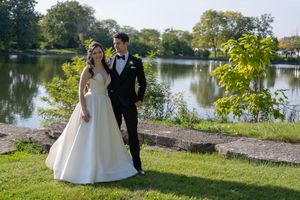Bride in white ball gown and groom in black tuxedo pose by a serene lake surrounded by lush greenery.