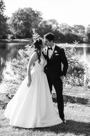 Black and white photo of a bride and groom sharing a kiss near a serene lake surrounded by lush trees.