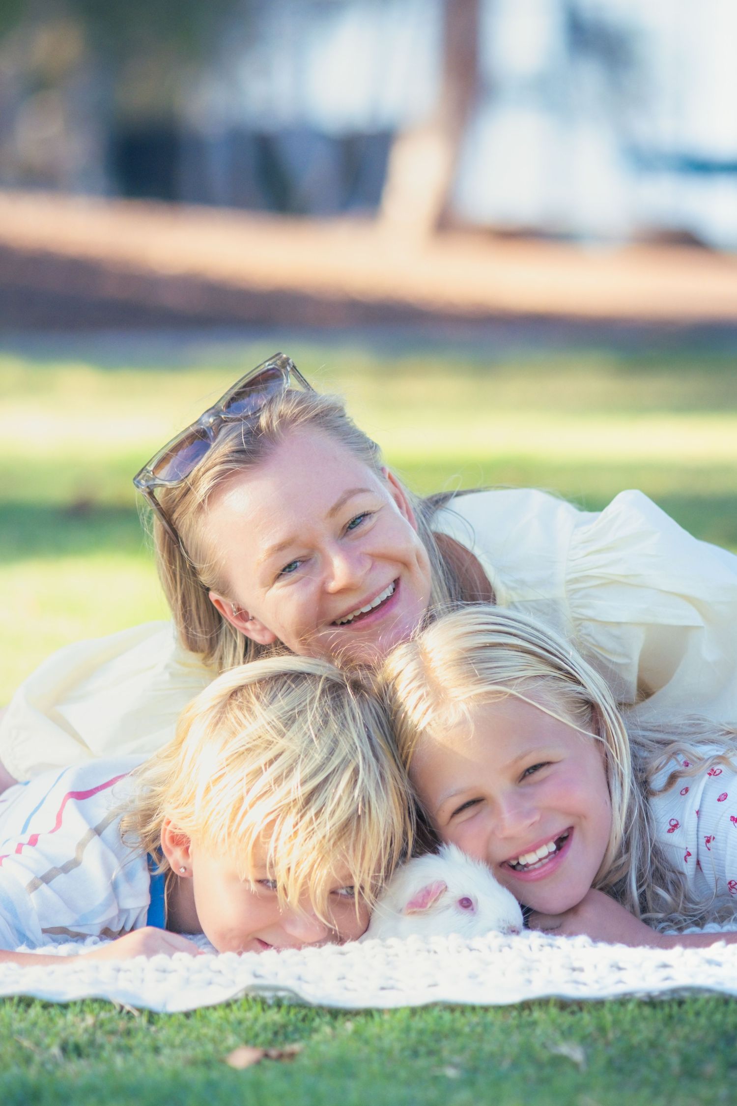A smiling mother and two blonde children lying on grass in a sunny park with a white guinea pig.